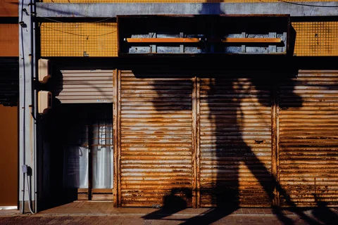 Long branching shadows cross rusty closed storefront shutters beneath dark signs and small yellow tiles.