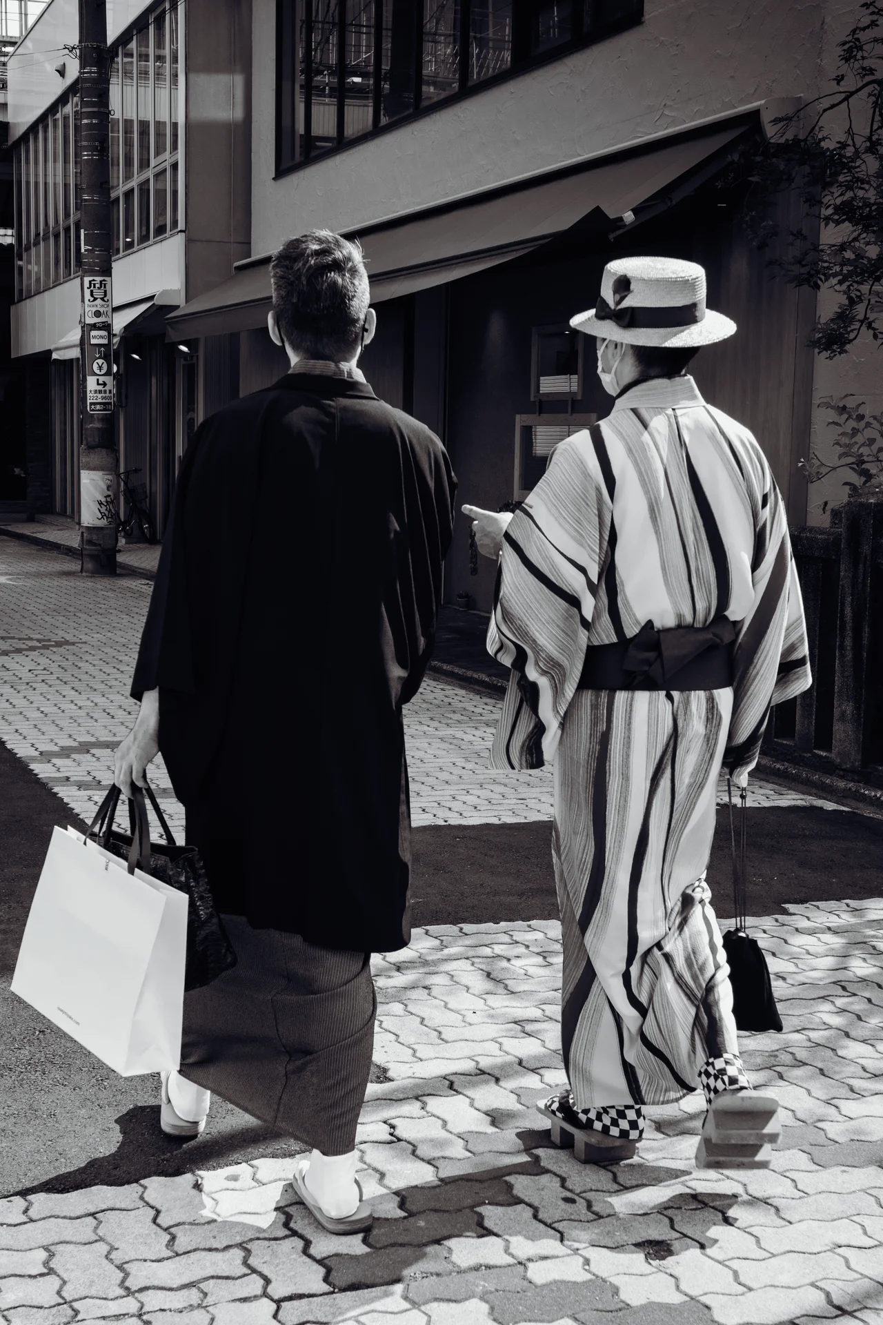 Two people walk away along a brick street, one wearing a striped robe and brimmed hat.