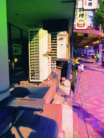 Outdoor air-conditioning units line a narrow storefront ledge beside a brightly colored sidewalk and projecting signs.