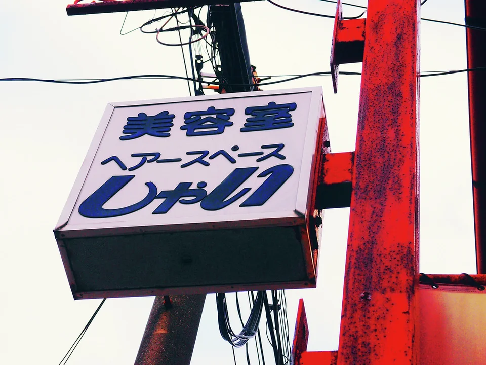 A pale projecting salon sign with blue lettering hangs beside a red metal support and utility cables.