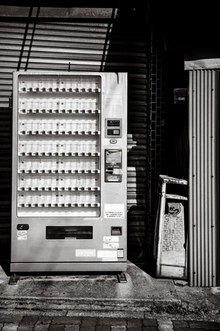 A tall vending machine stands before closed shutters beside a narrow dark opening in monochrome.