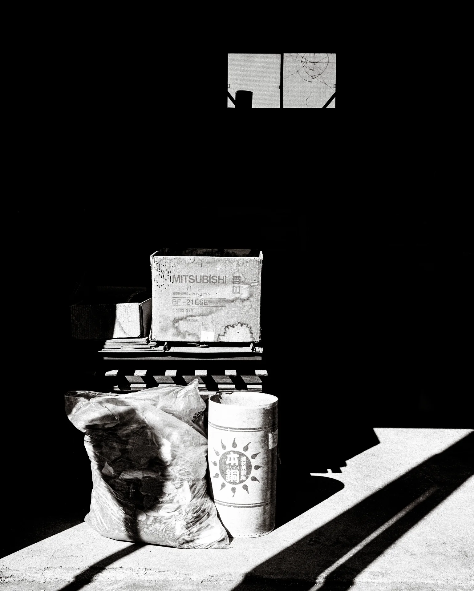 Bags and a bucket catch sunlight at the entrance of a dark workshop in black and white.