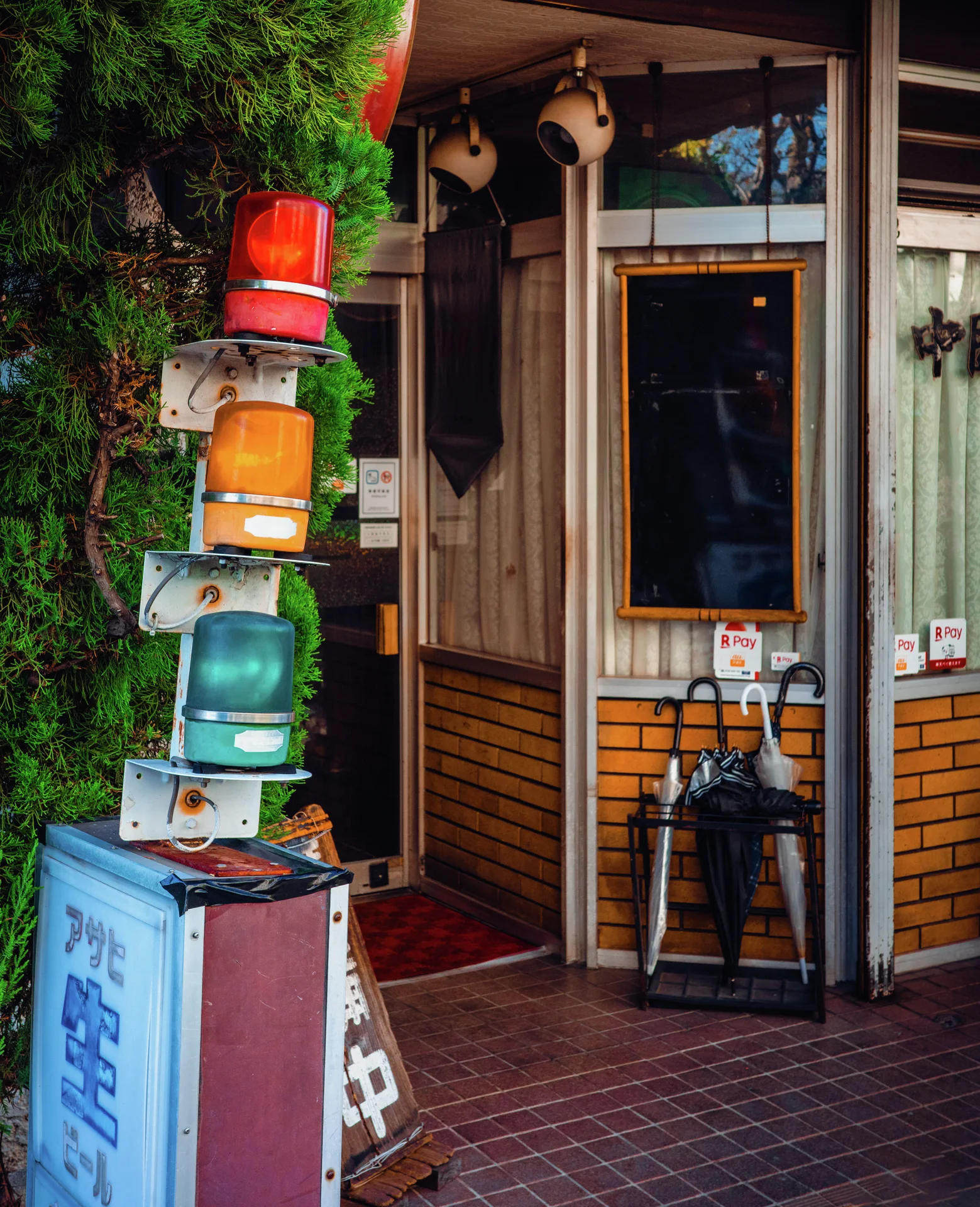 Stacked red, amber, and green lights stand beside a shop entrance with umbrellas and leafy greenery.