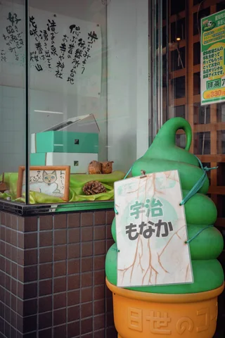 A large green soft-serve model stands in an orange cone beside a shop window and handwritten sign.