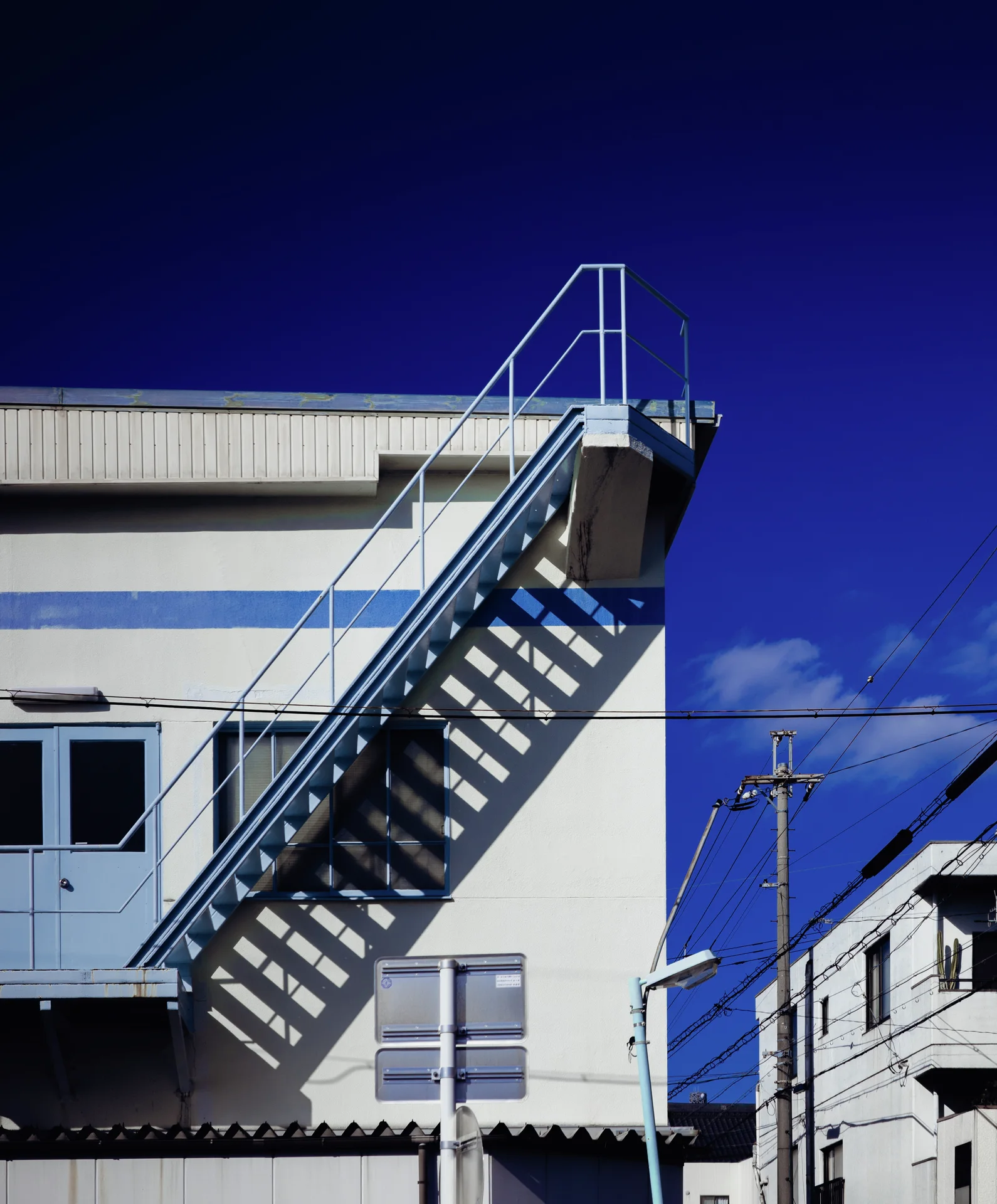 An exterior metal staircase climbs a pale building and casts striped shadows beneath deep blue sky.