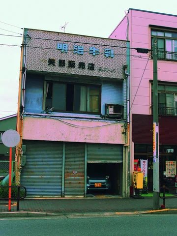 A narrow weathered shop has a closed shutter and shaded vehicle bay beside a taller pink building.