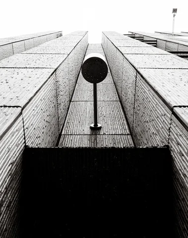 A circular dark fixture projects between towering brick-clad walls viewed upward in monochrome.