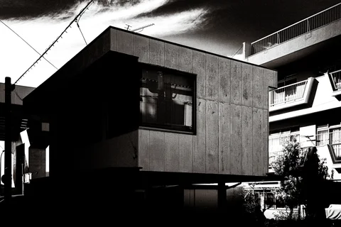 A boxy upper room projects over a dark building beside apartment balconies in monochrome.