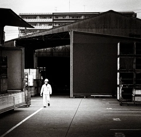 A person in pale work clothes and a hard hat crosses an industrial yard in black and white.