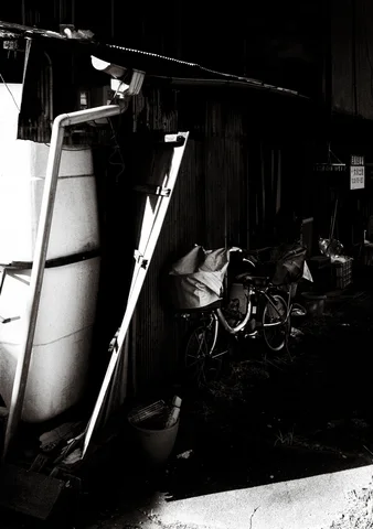 Two long ladders lean beside a large pale panel in a dark cluttered space in black and white.