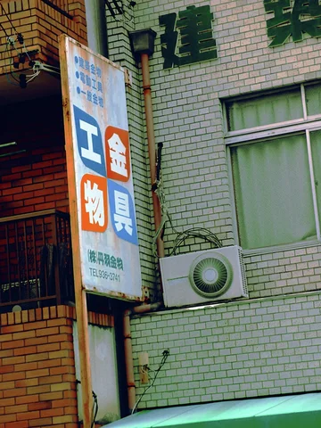 A narrow multicolored sign hangs beside a pale tiled facade with an air-conditioning unit and window.
