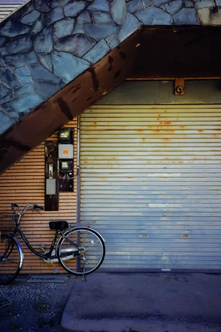 A bicycle rests before closed pale shutters beneath a steep stone-faced exterior staircase.