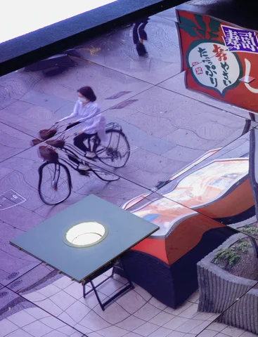 Two cyclists pass a sidewalk table and colorful street fixtures seen from above.