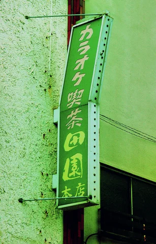 A slim green sign with pale Japanese characters projects from a cracked pale wall.