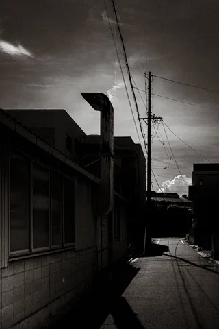 A curved vent pipe rises above a shadowed alley lined with low buildings in black and white.