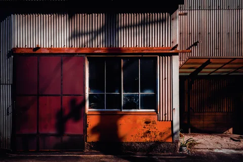 Strong shadows cross red doors and an orange lower wall beneath corrugated siding and dark windows.