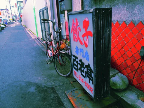 A tall restaurant sign stands beside a parked bicycle and red wall along a narrow sidewalk.