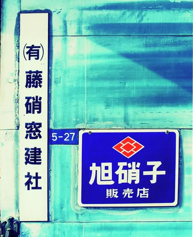 White vertical and blue rectangular signs hang on a turquoise-toned weathered exterior wall.