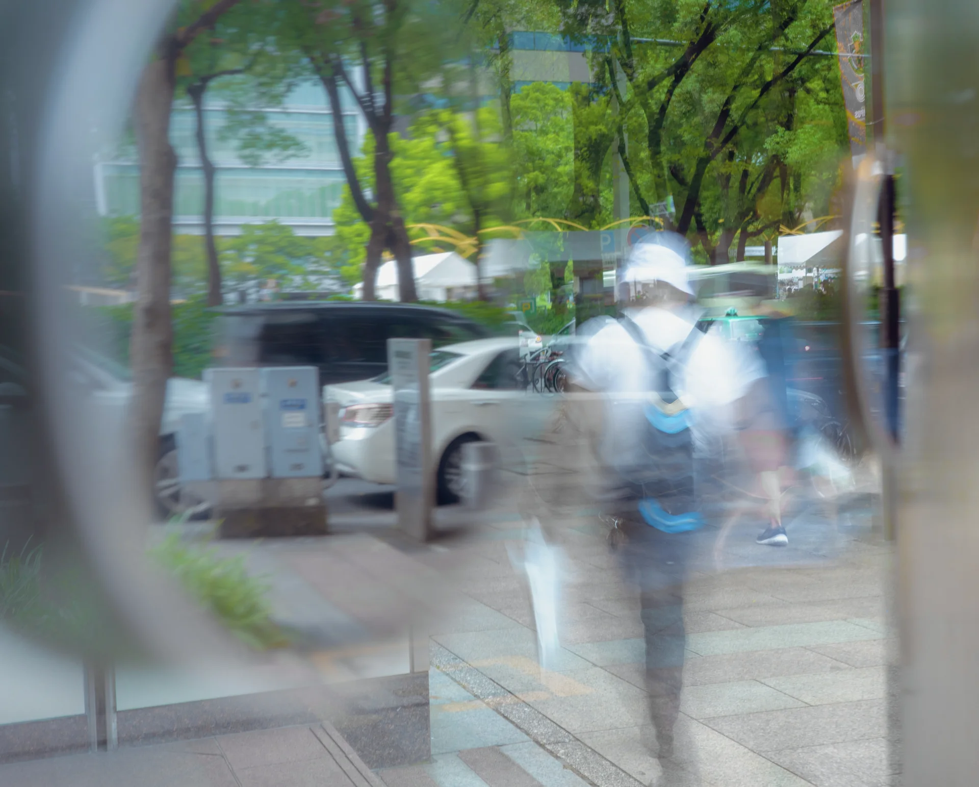 A blurred pedestrian with a blue backpack appears through glass, with parked cars and green trees behind.
