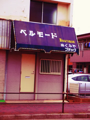 A purple boutique awning hangs above a narrow pale door and barred window beside a parked white car.