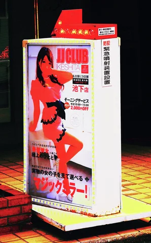 A freestanding illuminated sign displays a woman in red clothing and Japanese lettering beside a dark entrance.