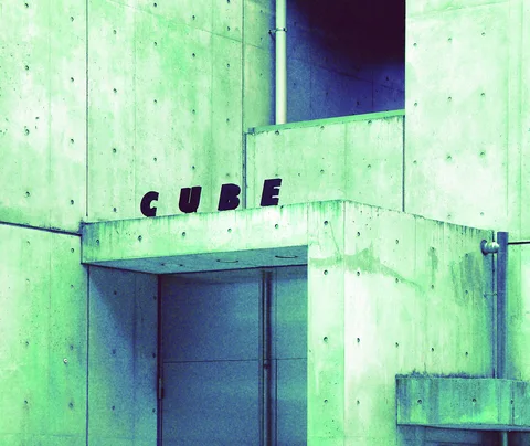 Dark letters spelling Cube sit above a recessed doorway in a pale green concrete facade.