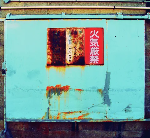 Rust stains run down turquoise metal doors beneath two small rectangular warning signs.