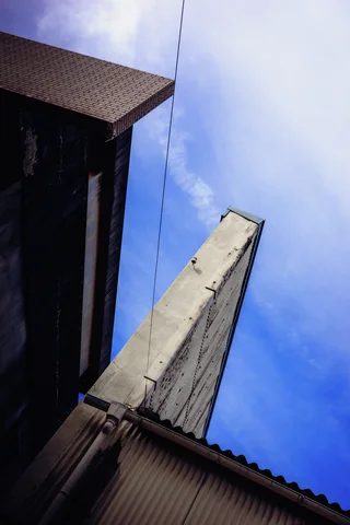 A tall narrow concrete structure rises between dark building edges and corrugated roofing against blue clouds.