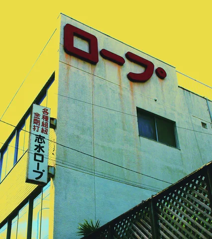 Dark red Japanese lettering tops a pale rust-streaked building beside a vertical sign under yellow sky.