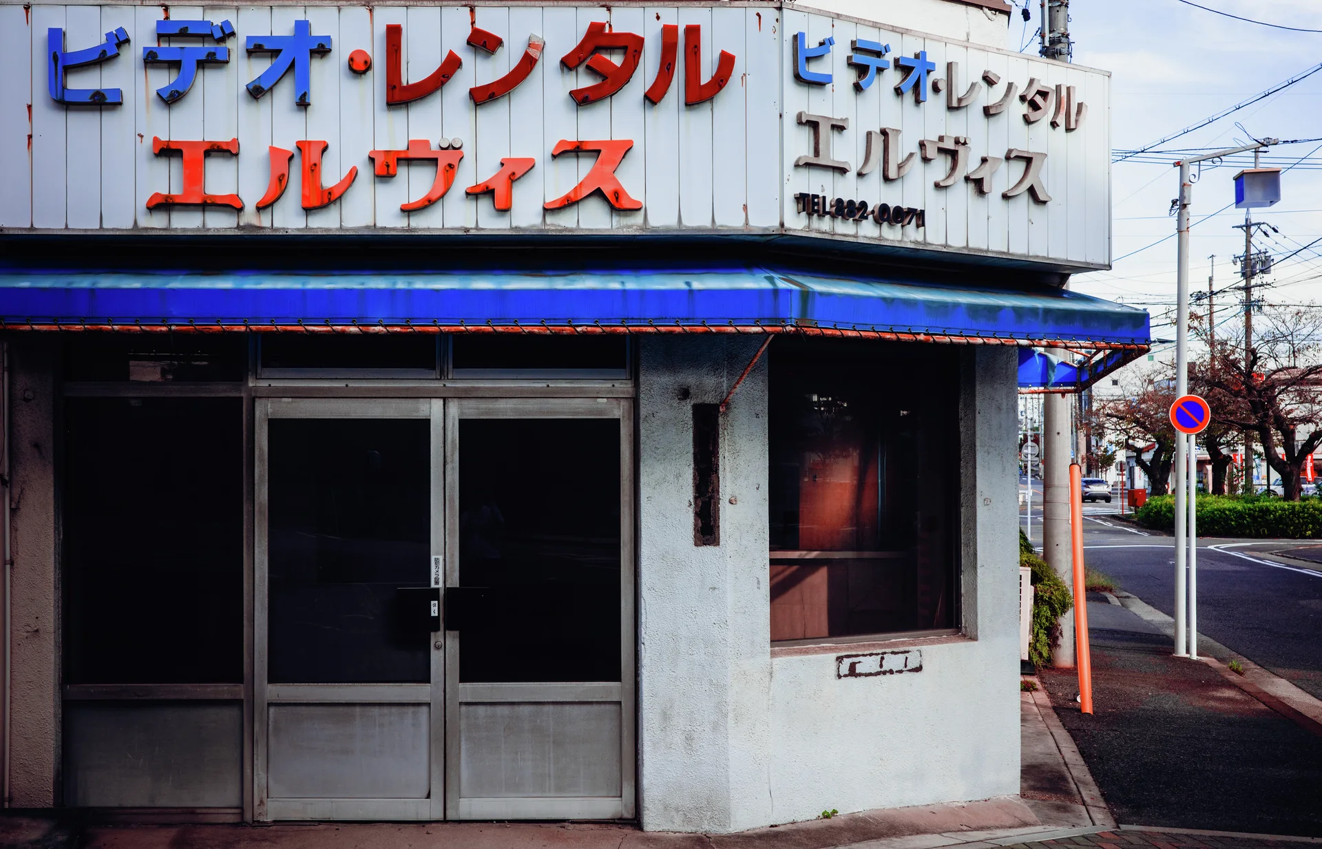 Red and blue Japanese lettering crowns a closed corner storefront beneath a blue awning.