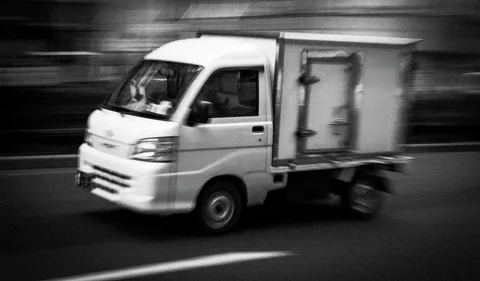 A small white box truck crosses a horizontally blurred street in a black and white view.