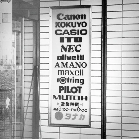 A tall rectangular sign lists electronics and stationery brands beside a tiled wall in black and white.
