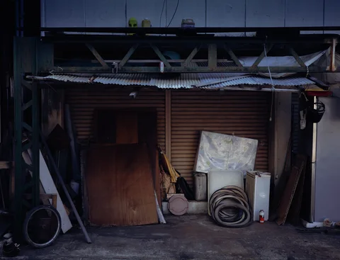 Boards, coiled tubing, and metal panels lean against closed shutters beneath a sagging canopy.