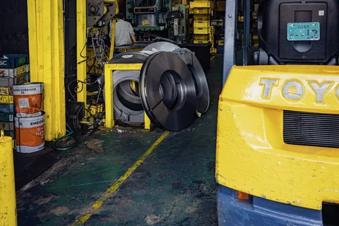 Yellow industrial machinery surrounds a large dark metal coil inside a cluttered workshop.