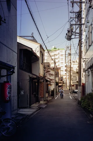 A narrow alley extends between mixed-height buildings beneath numerous utility wires toward a distant pedestrian.