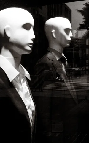 Two pale bald mannequins wear collared shirts and dark jackets behind reflective glass in black and white.