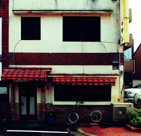 A bicycle stands outside a weathered pale building with dark windows and two red grid-patterned awnings.