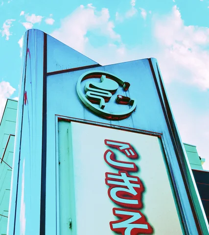 A pale blue angular sign carries raised Japanese lettering and a circular emblem beneath bright clouds.
