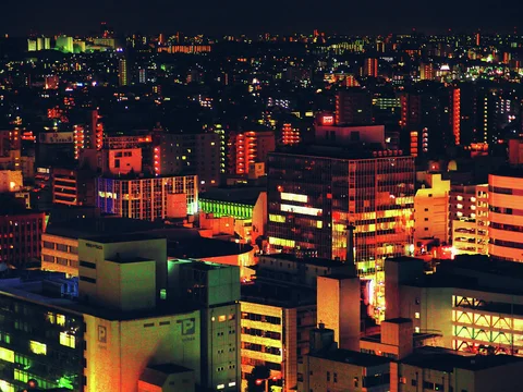 Colorful illuminated windows and signs dot a dense cityscape beneath a dark sky.