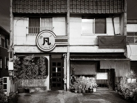 A two-story tiled-roof building has a circular sign and potted plants by its entrance in black and white.