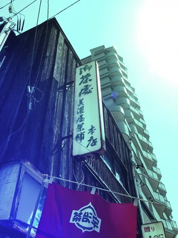 A tall narrow sign hangs on a weathered wooden building beside a high-rise with stacked balconies.