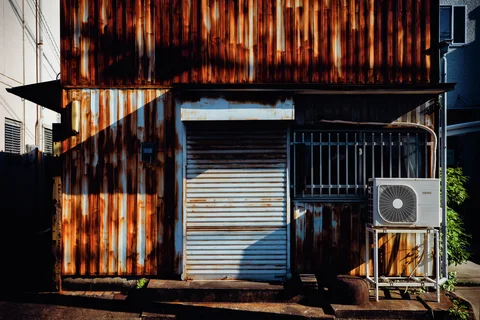 Rust covers a corrugated building facade around a closed shutter, barred window, and air-conditioning unit.