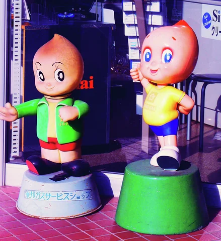 Two smiling cartoon child statues wear green and yellow outfits on round bases outside a shop.