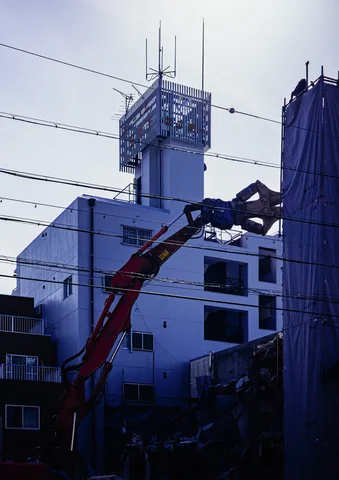 An orange excavator arm reaches beside a blue-toned building with a narrow rooftop tower and antennas.