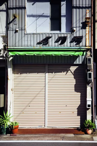 Closed pale shutters sit beneath a green awning and raised lettering on a corrugated metal storefront.