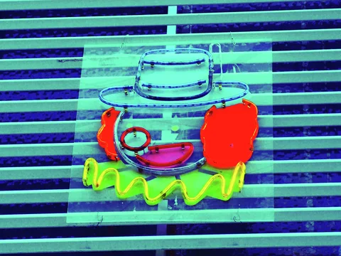 A colorful neon clown face with red hair and a small hat hangs before horizontal turquoise blinds.