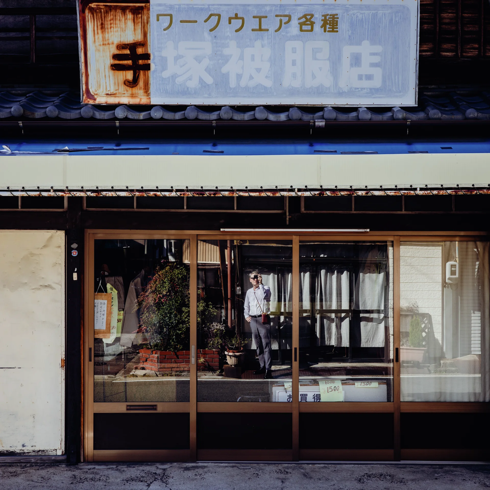 A person stands behind glass shop doors beneath a faded sign and a narrow blue roof edge.