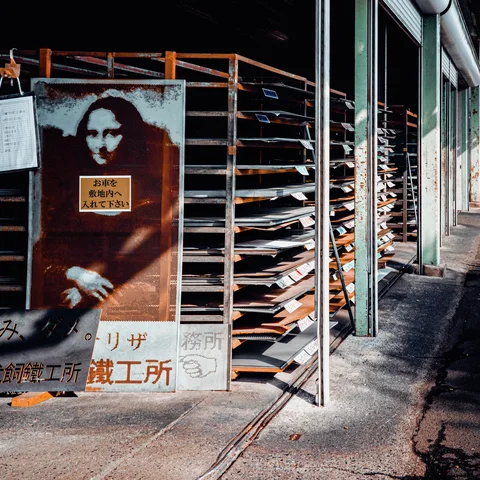 A reproduction of the Mona Lisa leans beside tall racks of flat materials outside a workshop.
