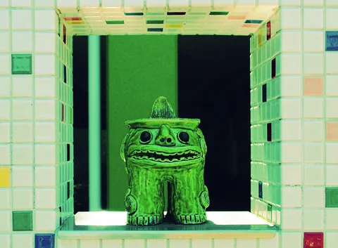 A small green creature sculpture with round eyes sits in a square recess framed by multicolored tiles.