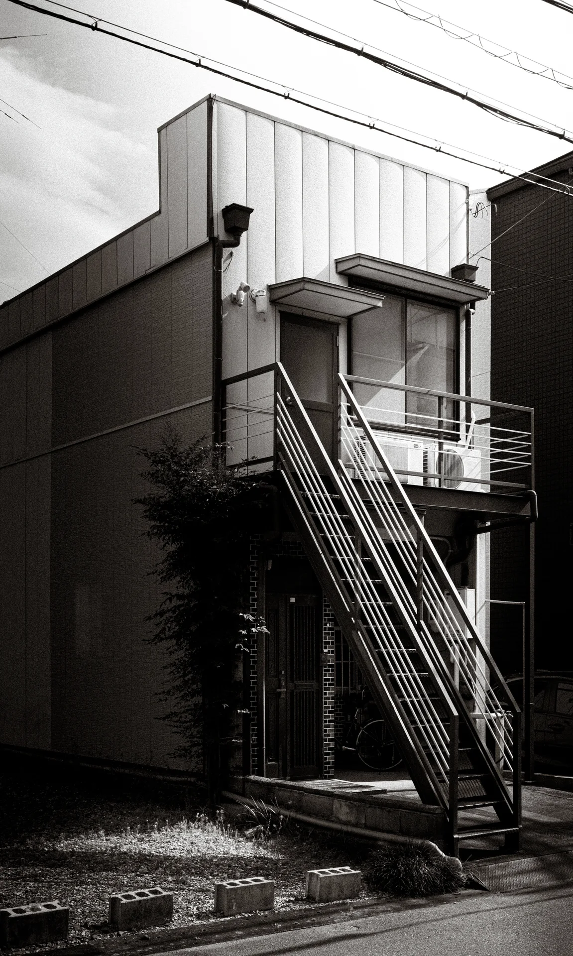 A steep exterior metal staircase climbs a narrow building beside a grassy vacant plot in black and white.
